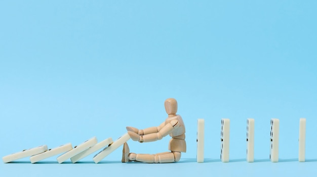 A wooden puppet toy holds back a falling domino on a blue background representing the concept of a strong personality