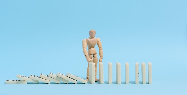 A wooden puppet toy holds back a falling domino on a blue background representing the concept of a strong personality