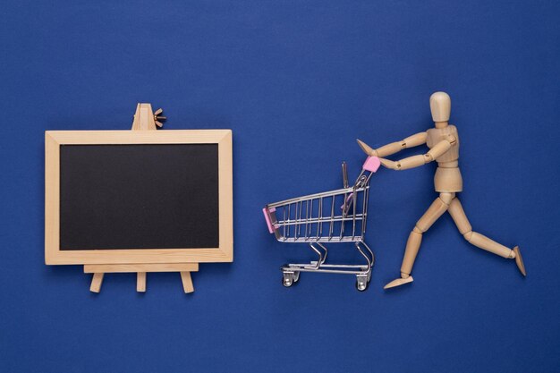 Wooden puppet with shopping trolley and Mini Empty chalk board with copy space on blue background Shopping sale concept