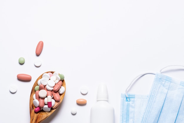 Wooden spoon full of medical capsules and pills, nasal spray and protective facial masks on white background.