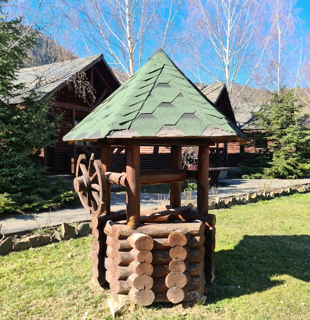 A wooden structure with a green roof is in front of a building with a log cabin in the background