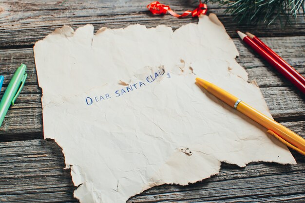 On the wooden table are an old yellow paper that says, Dear Santa Claus red, pencils, pens assorted colors gift and tree branches for the new year