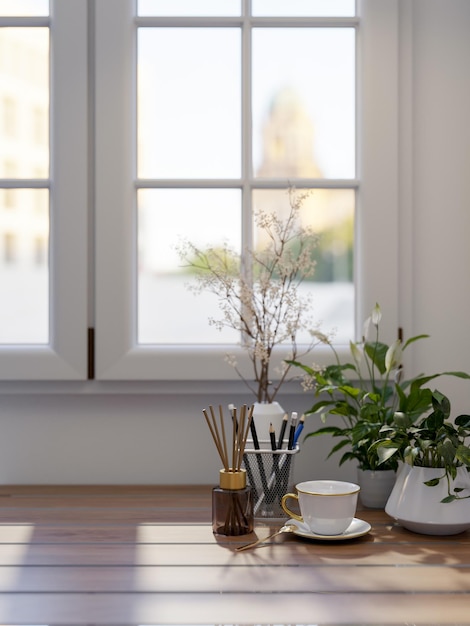 A wooden table by the window in a room features a coffee cup a reed diffuser and decorative plants