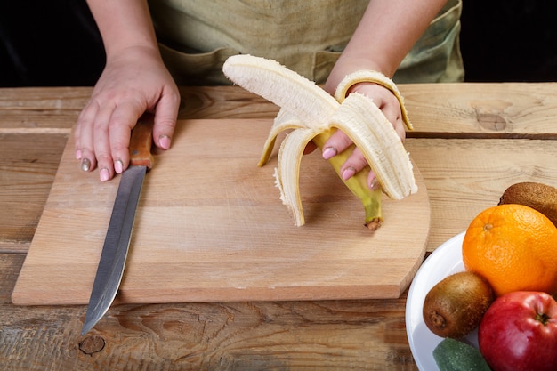 On a wooden table on a cutting board, a woman removes the skin from a banana