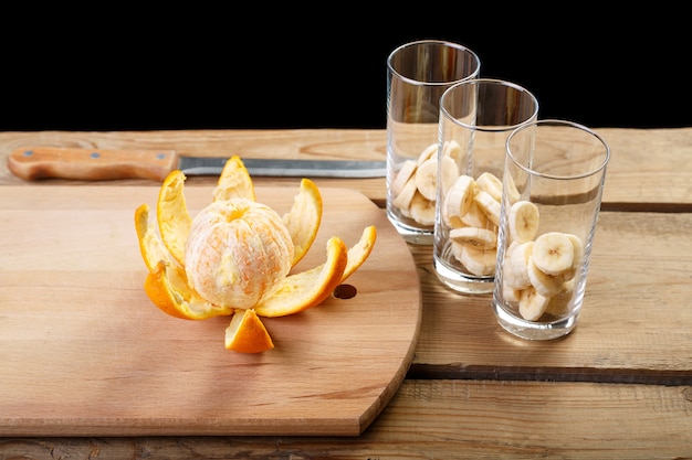 On a wooden table there is a peeled orange and three glasses with bananas for a cocktail