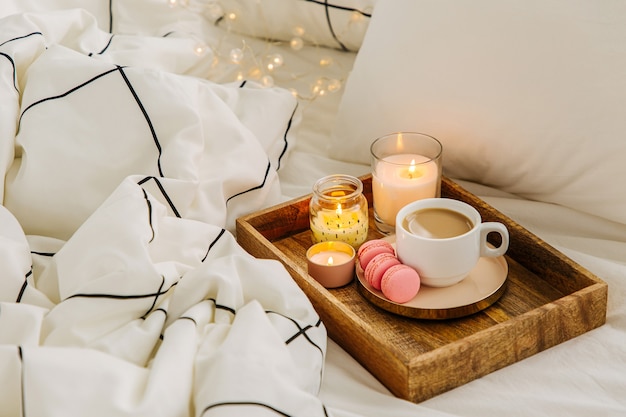 Wooden tray of coffee and candles on bed. White bedding sheets with striped blanket and pillow. Breakfast in bed. Hygge concept.