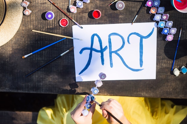 The word art is written in blue paint on a white sheet of paper among the brushes and paints for drawing