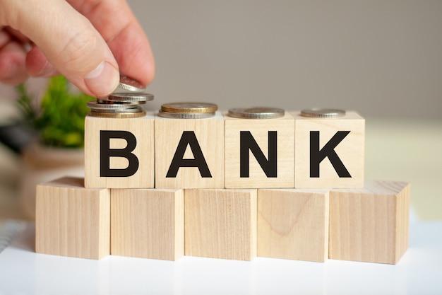 The word bank written on wood cubes. A man's hand places the coins on the surface of the cube. Green potted plant on the background. Business and finance concept.