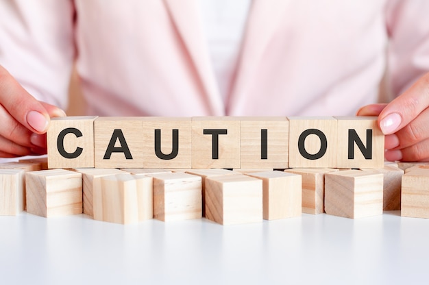 Word caution is written on wooden cubes standing on the white surface of the table on the pink background