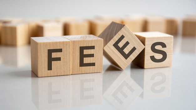 Word FEES on wooden cubes, gray background. Reflection on the mirrored surface of the table. Selective focus.