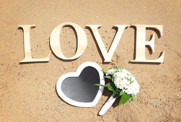 Photo word love composed of wooden letters beautiful bouquet and chalkboard in shape of heart on sand beach wedding concept