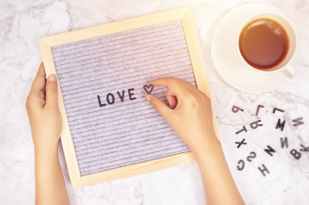 Word LOVE on letter board with woman's hand holding heart symbol on white marble desk 