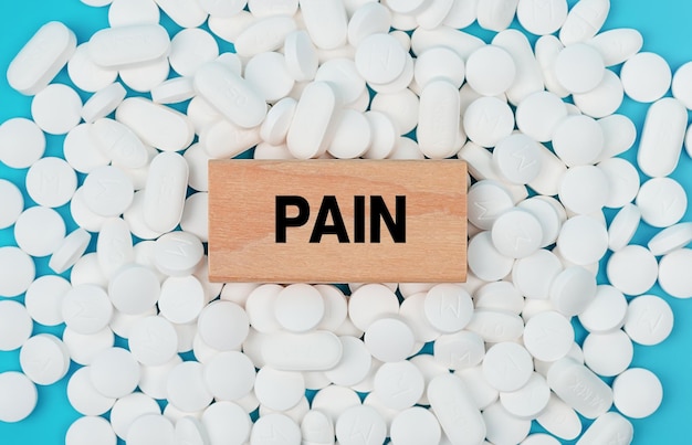 The word Pain is written on wooden blocks on pile of white pills on blue background. pain medication