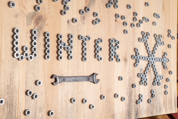 Word winter and a snowflake are lined with metal nuts on a wooden table next to a wrench