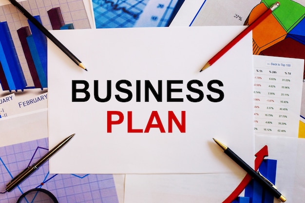 The words BUSINESS PLAN is written on a white background near colored graphs, pens and pencils