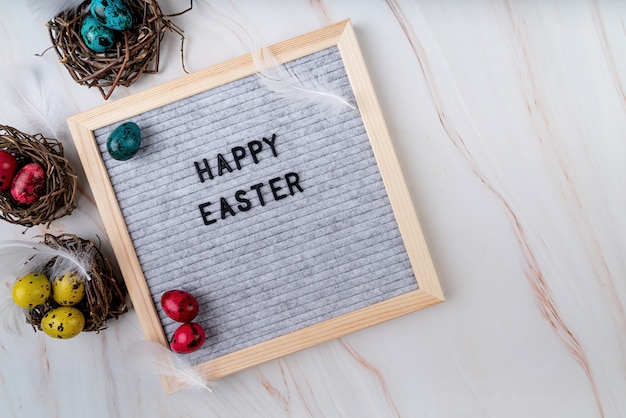 The words Happy Easter on grey felt letter board decorated with quail and chicken eggs and feathers on marble