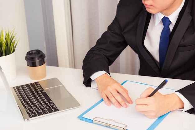 Work from home, Asian young businessman smile wearing suit video conference call or facetime by laptop computer on desk and write taking notes on paper at home office