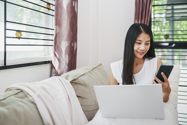 Work from home and online shopping concept, Happy business asian woman using mobile phone and laptop computer with notebook on sofa in living room at home