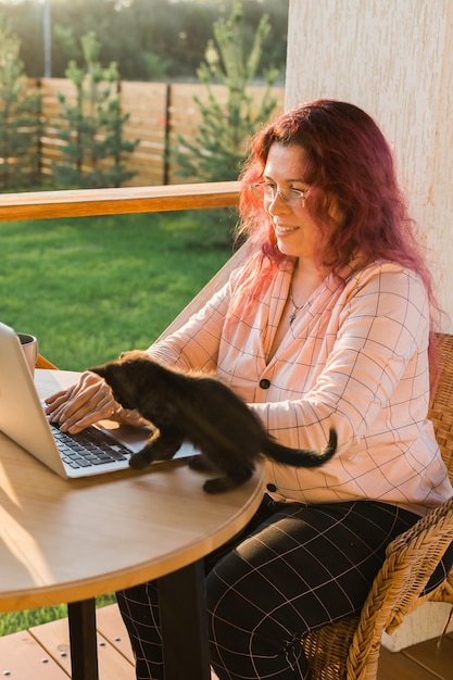 Work from home or study online and videocall with a woman gardener working at laptop outdoor on terrace with kitten closeup sitting on ratang chair around houseplant and flower pots