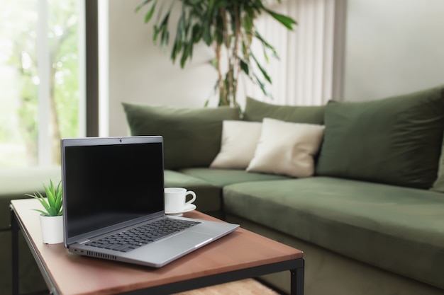 Work from home, workspace, desktop, remote work concept, gray thin laptop computer black empty screen on brown wooden table with white cup of coffee, green sofa, flowerpot. Stylish apartments