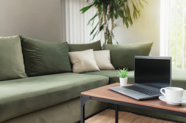 Work from home, workspace, desktop, remote work concept, gray thin laptop computer with black empty screen on brown wooden table with white cup of coffee, green sofa, flowerpot. Apartment comfort zone