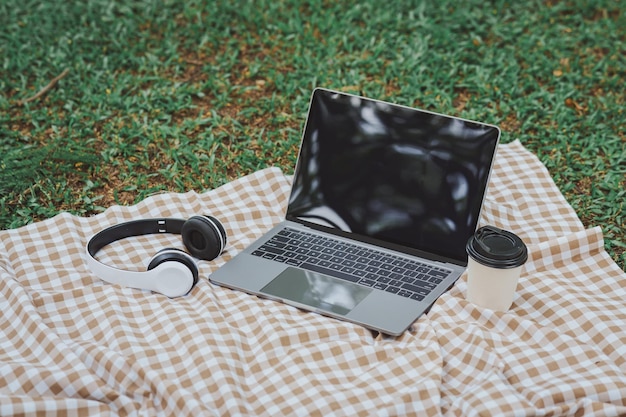 Work on laptop on picnic blanket on the lawn in the park next to a headphone notebook and cup of hot coffee Freelancer work concept Top view flat lay Work form anywhere concept