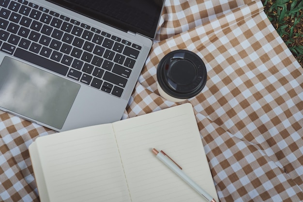 Work on laptop on picnic blanket on the lawn in the park next to a headphone notebook and cup of hot coffee Freelancer work concept Top view flat lay Work form anywhere concept