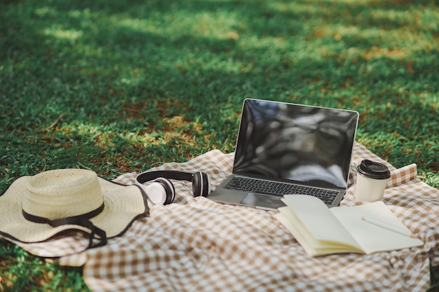 Work on laptop on picnic blanket on the lawn in the park next to a headphone notebook pen hat and cup of hot coffee Freelancer work concept Top view flat lay Work form anywhere concept