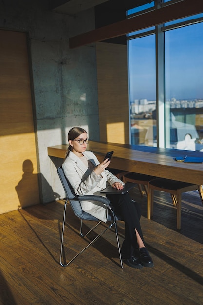 Work in a modern office Positive young businesswoman in casual clothes and glasses sitting in the office with a mobile phone while working on a remote project using a laptop