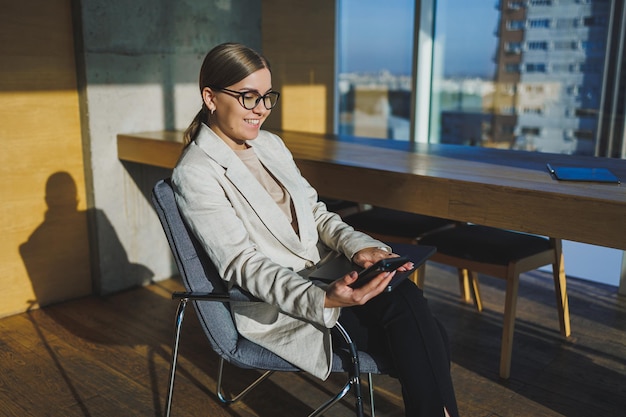 Work in a modern office Positive young businesswoman in casual clothes and glasses sitting in the office with a mobile phone while working on a remote project using a laptop