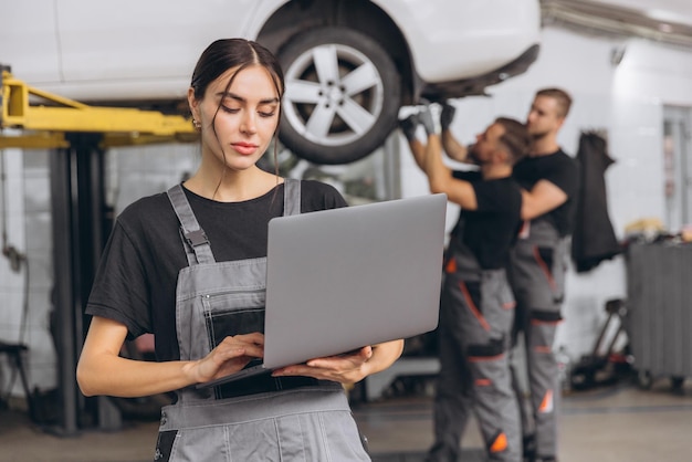 Photo worker of autoservice beautiful young woman holding laptop in hands and looking at camera smiling mechanics fixing repairing automobile lifted on bridge behind