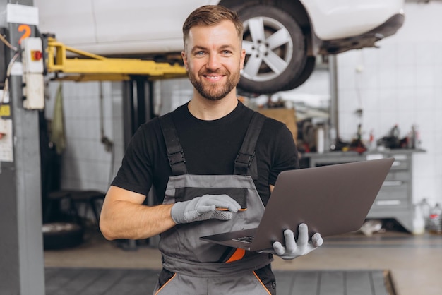 Worker of autoservice smiling bearded man holding laptop in hands Mechanic fixing repairing automobile lifted behind