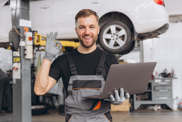 Photo worker of autoservice smiling bearded man holding laptop in hands and showing thumbs up mechanic fixing repairing automobile lifted behind