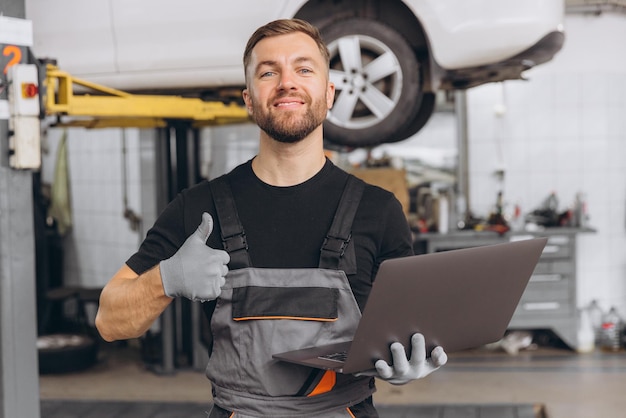 Worker of autoservice smiling bearded man holding laptop in hands and showing thumbs up Mechanic fixing repairing automobile lifted behind