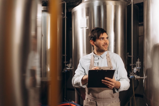 Worker brewer business owner works on factory with equipment modern tech Satisfied smiling young attractive guy in an apron holds tablet and checks metal boilers at plant in brewery interior