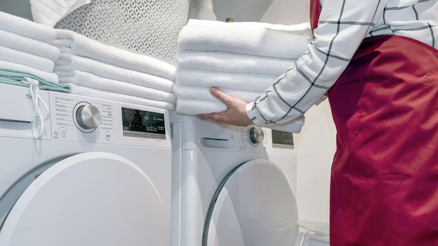 Worker in the hotel holds a lot of towels in hands near Dry machine in the laundry