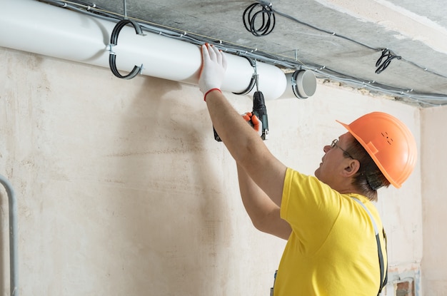 The worker is fixing ventilation pipes to the ceiling with a screwdriver