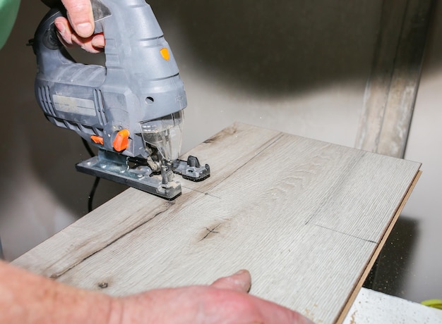 Worker is holding in hands power jigsaw and cutting laminate flooring in apartment Maintenance repair works renovation with scroll saw Wooden parquet floor planks indoors