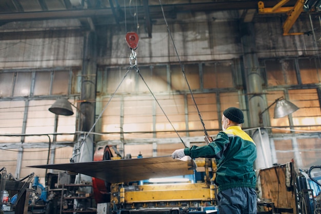 Worker lift metal sheet with crane chain hoist with remote controller and hook at industrial factory plant