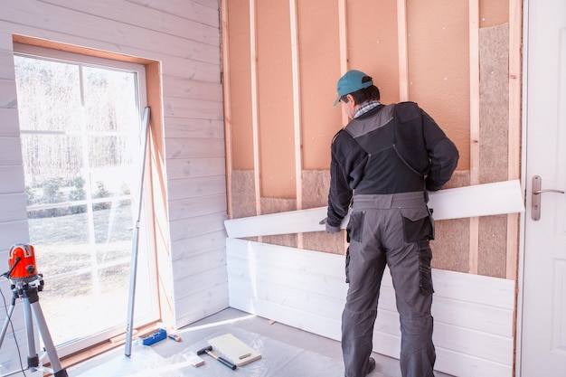 The worker makes finishing works of walls with a white wooden board using laser line level Building heatinsulating ecowooden frame house with wood fiber plates