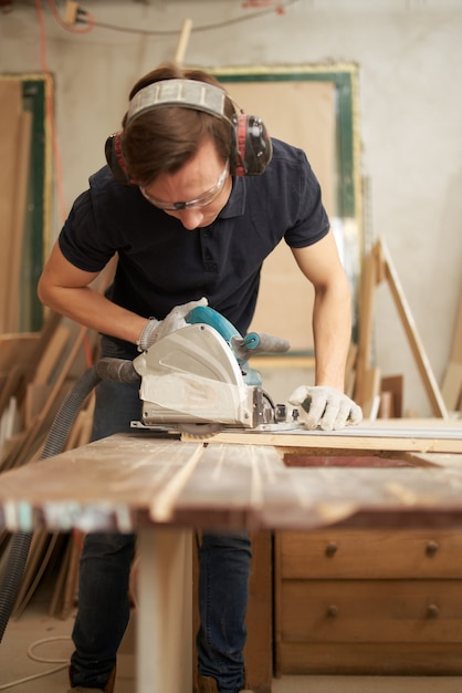 Worker male in safety glasses and white gloves works on jigsaw in workshop