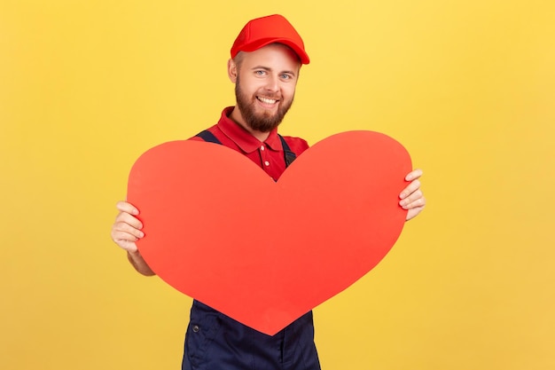 Worker man wearing overalls and red cap holding big red heart fast gifts delivery during holidays