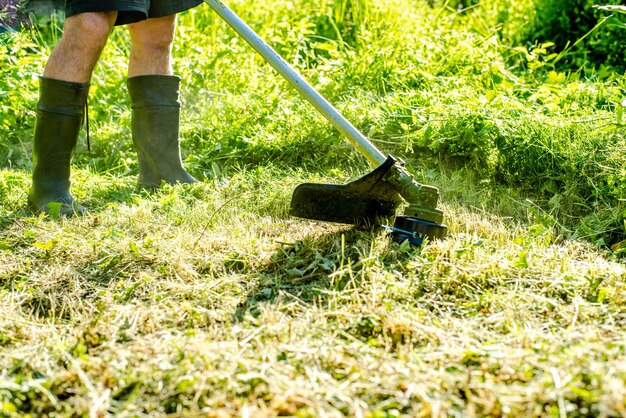 Worker mowing tall grass with electric or petrol lawn trimmer in city park or backyard Gardening care tools and equipment Process of lawn trimming with hand mower