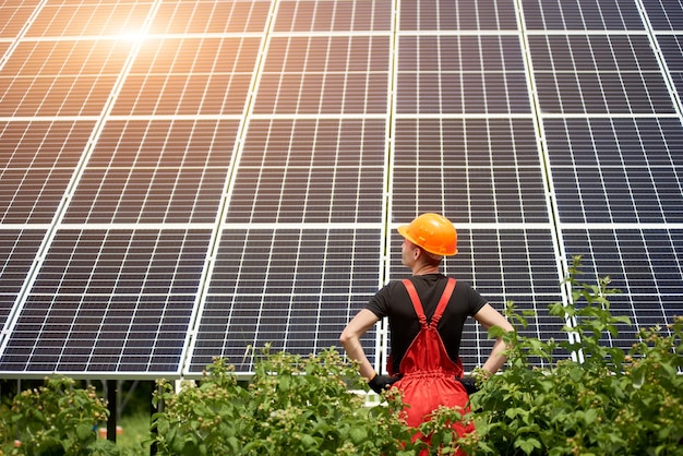 Worker in orange uniform stands with his back to camera against the background of large plantation solar batteries