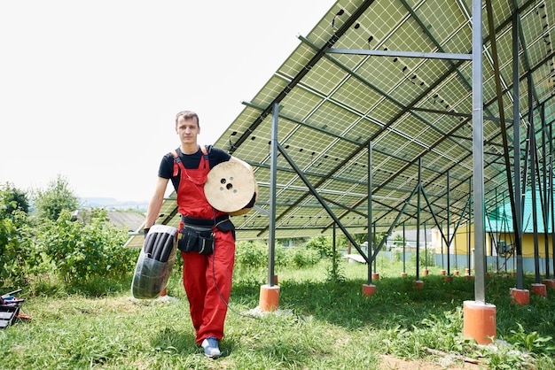 Worker in an orange uniform with a coil of wires in his hands on a background of solar panels near the house