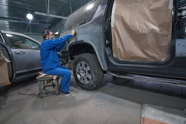 Worker in protective clothes sitting near car and doing grinding matting car bodies before painting and primer