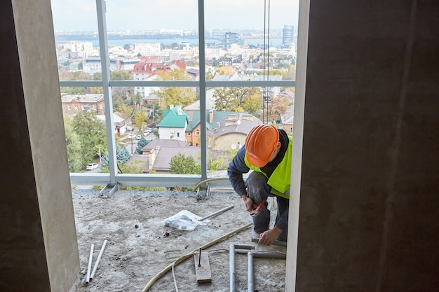 Worker in protective clothing and safety helmet installing plastic pipes using modern tools in flat of building under construction