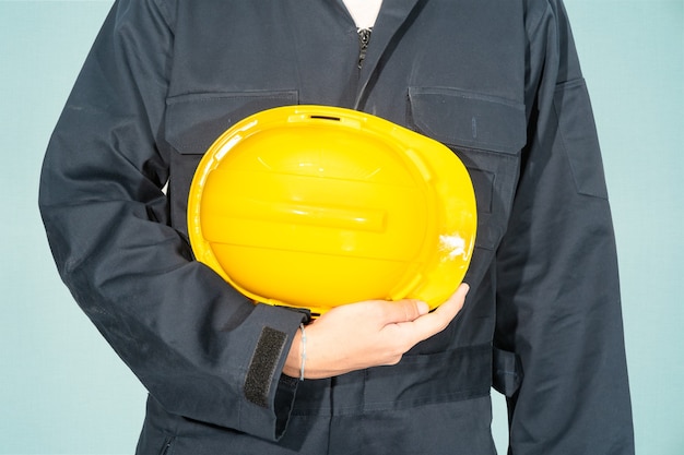 Worker standing in blue coverall holding yellow hardhat isolated on blue background