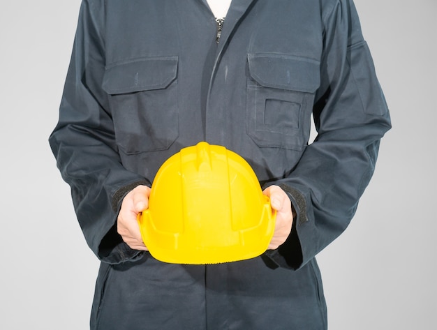 Photo worker standing in blue coverall holding yellow hardhat isolated on gray background