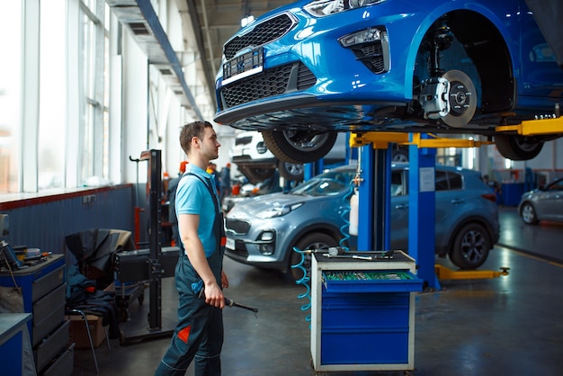 Worker in uniform checks vehicle bottom on lift, car service station. Automobile checking and inspection, professional diagnostics and repair